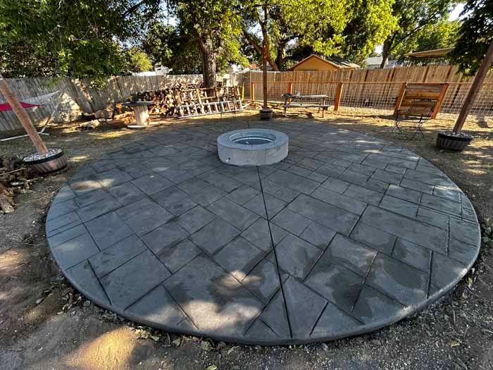 Circular concrete patio with fire pit, surrounded by a yard with trees and wooden structures.