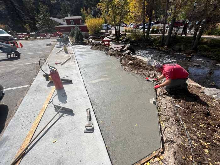 Person smoothing wet concrete near a creek. Construction materials and tools are visible. Sunny day.