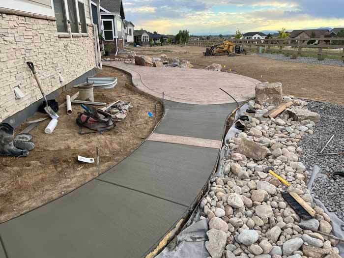 Newly poured concrete walkway and patio under construction beside a house, surrounded by rocks and dirt.