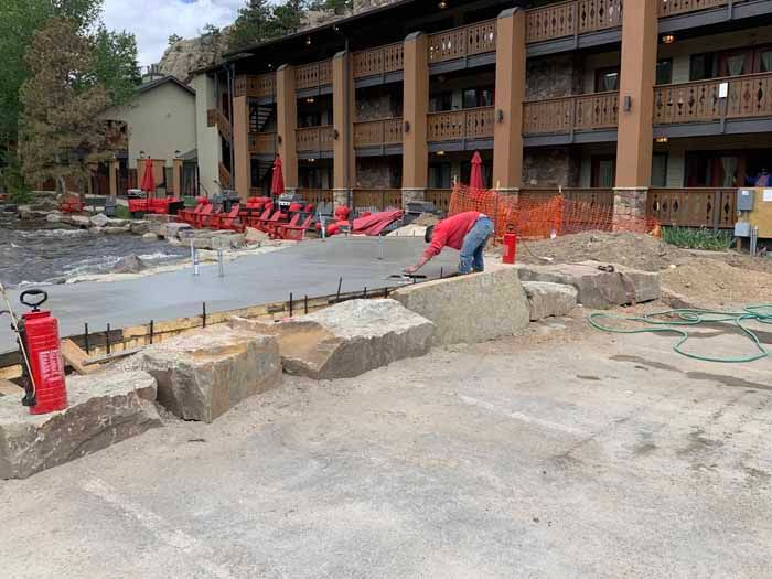Construction worker smoothing wet concrete near a building with red umbrellas and large rocks.