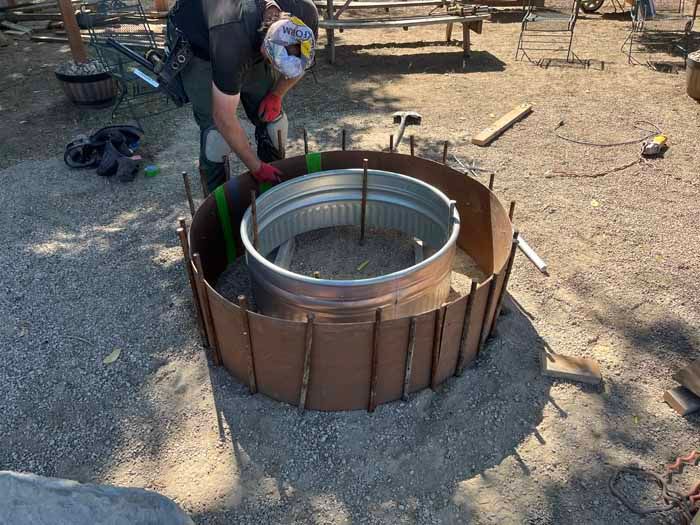 A person building a fire pit using a metal tub, surrounded by a ring of wood and rebar, outdoors.