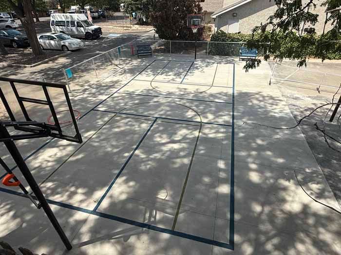 Basketball court with blue lines, hoop, and vehicles in the background.