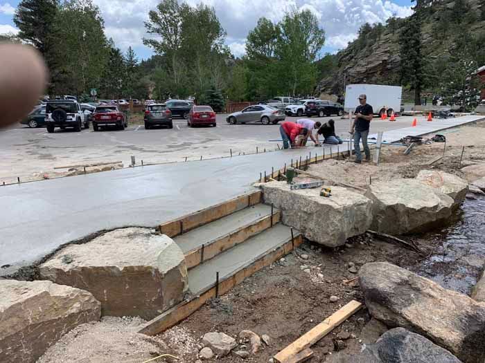 Concrete path and steps under construction near a parking lot, with workers, large rocks, and a stream.