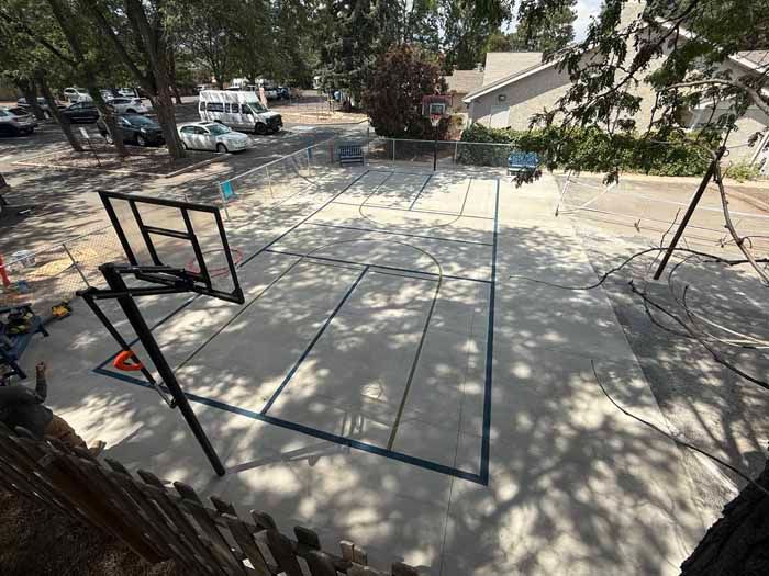 Basketball court, gray concrete with blue lines, with a black hoop, viewed from above, surrounded by trees and a building.
