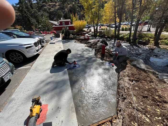 Men pouring concrete sidewalk next to a parking area and a small stream in a park.