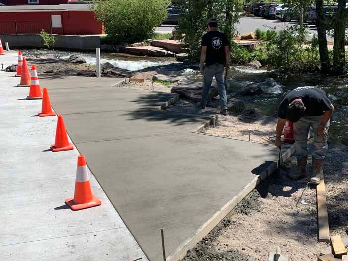 Workers pouring concrete on a walkway near a stream; orange cones line the edge.