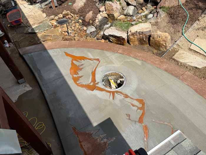 Concrete patio with orange stains near a circular drain, bordered by a reddish path and rocks.