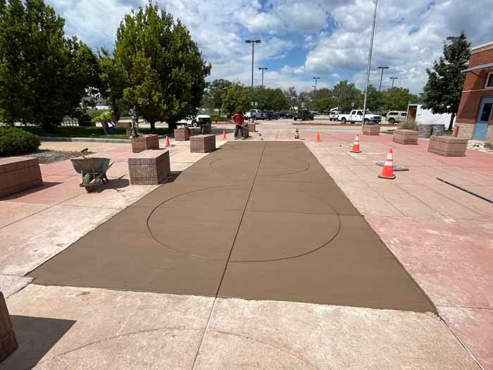 Newly poured concrete basketball court; worker in red shirt, orange cones, construction site.
