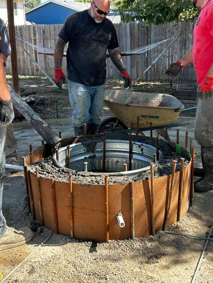 Men pouring concrete into a circular form. Construction site outdoors; brown, gray and green tones.
