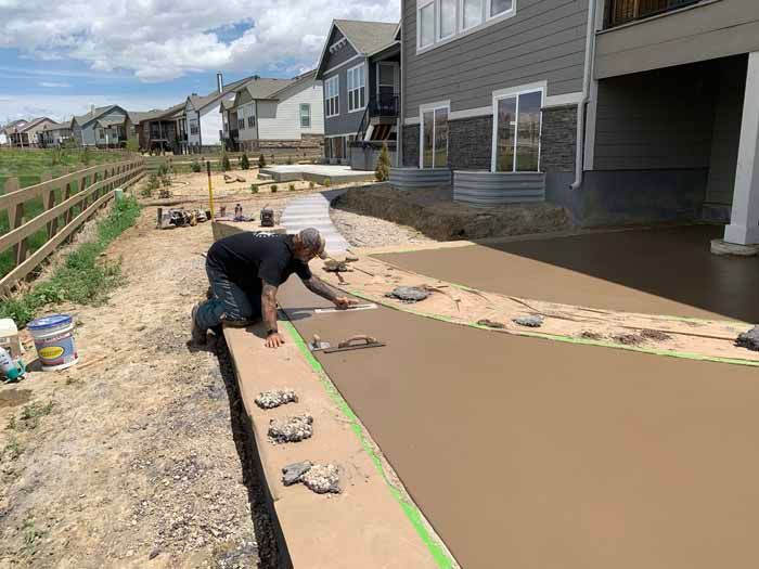 Man smoothing wet concrete walkway with decorative rocks and houses in background.