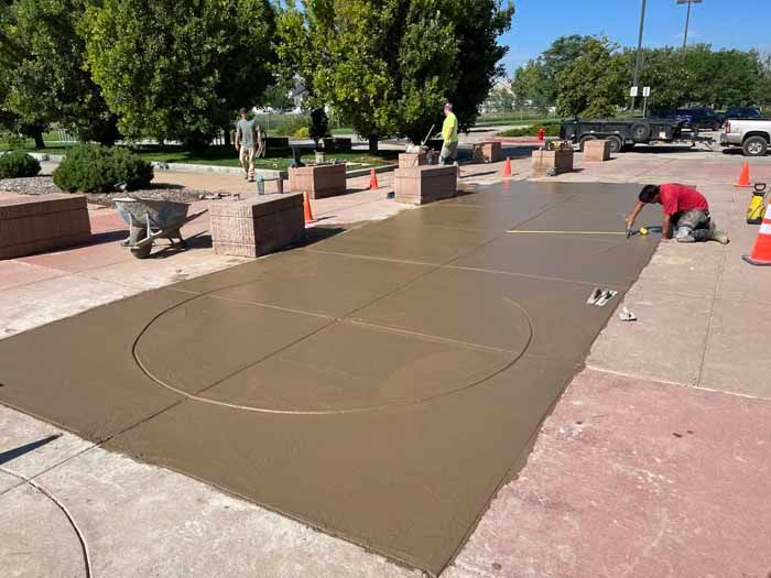 Workers constructing a concrete pad with a basketball court outline outdoors on a sunny day.