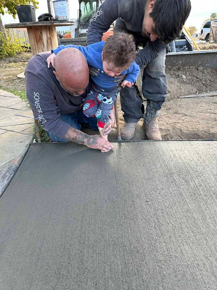 Two people help a child imprint their hand in wet concrete outdoors.