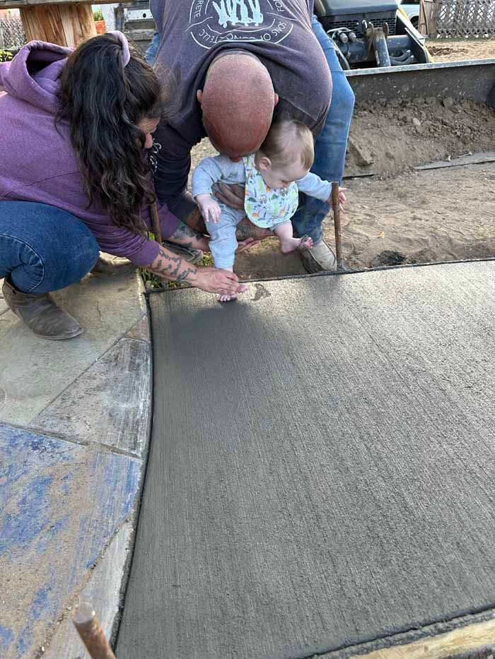 Family helping a baby stand on fresh concrete outdoors.