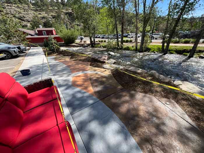 Exterior scene with a red couch on a sidewalk, next to a concrete path with decorative colored designs, and a flowing creek.