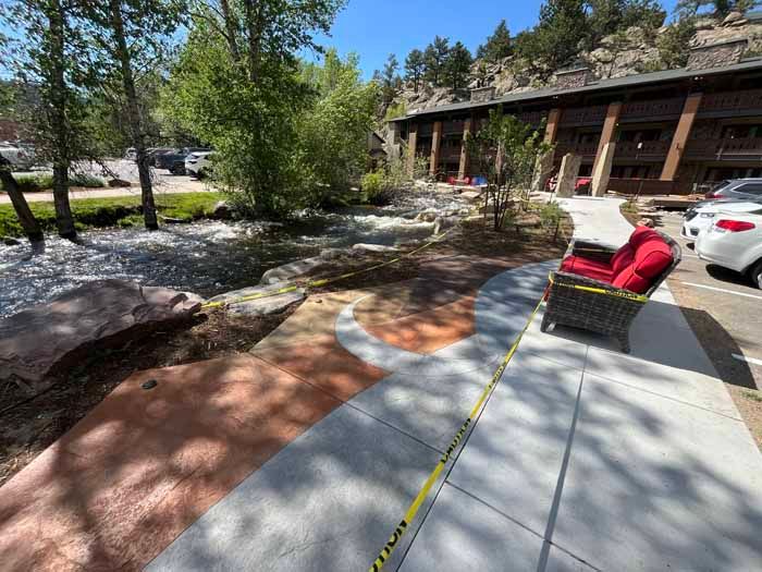 Sidewalk beside a stream and building, with outdoor seating. Yellow caution tape in foreground. Sunny day.