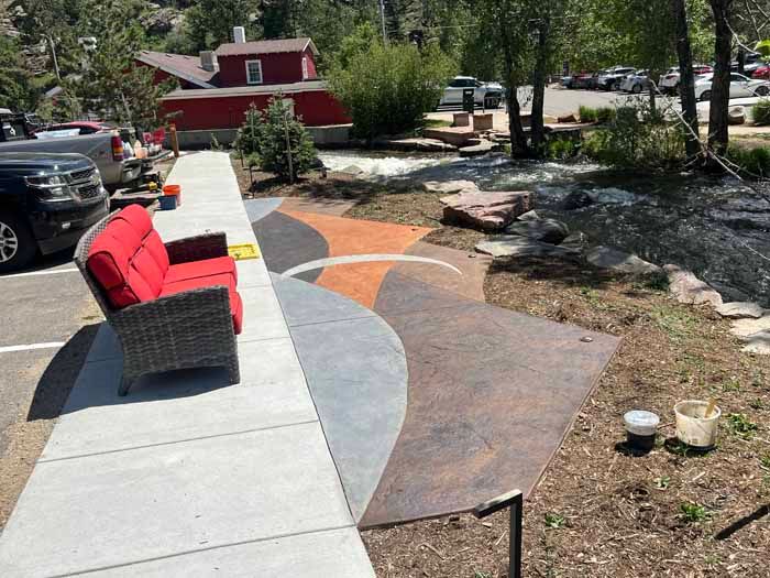 Outdoor seating area with red sofa, multi-colored pathway, next to a river. A red building is in the background.