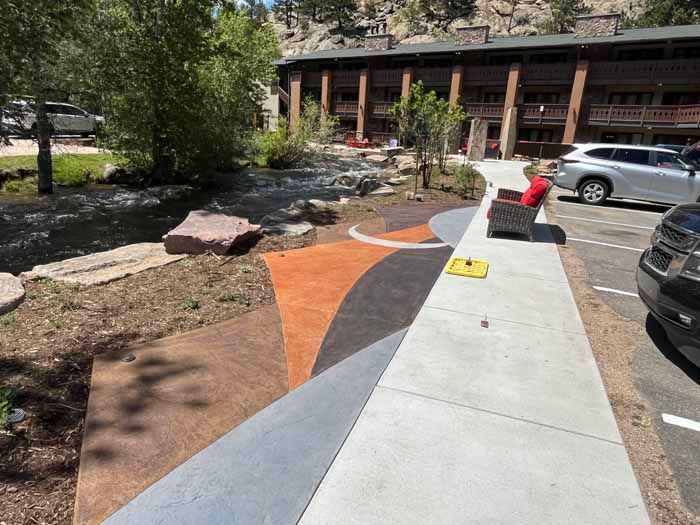 Sidewalk with colorfully stained concrete curves next to a creek and building. Parking area visible.