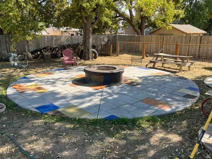 Circular patio with fire pit, colorful tiles, picnic table, and fence in a backyard setting.
