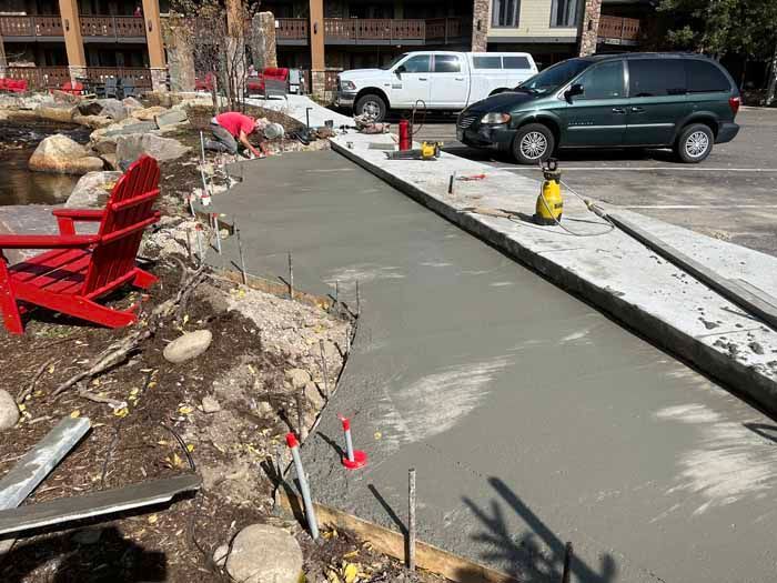 Concrete walkway being poured near a pond with red Adirondack chairs. Construction workers and vehicles visible.
