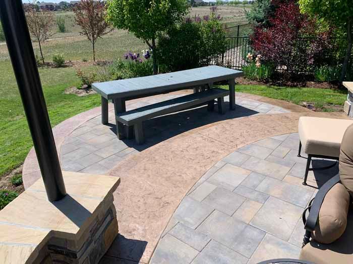 Patio with gray table and bench on a brick pathway, surrounded by green grass and plants.
