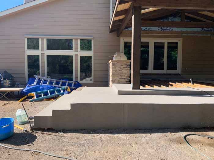 Concrete patio construction next to a house with windows and a covered patio, tools and materials scattered around.