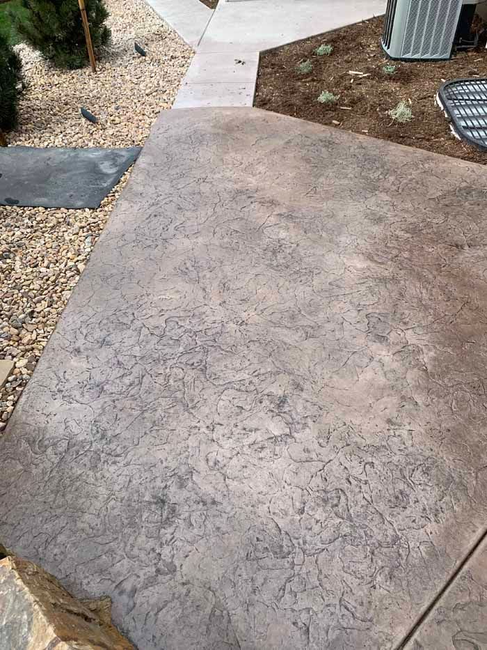 Textured brown concrete patio leading to a walkway. Gravel and plants on the left, an air conditioner is on the right.