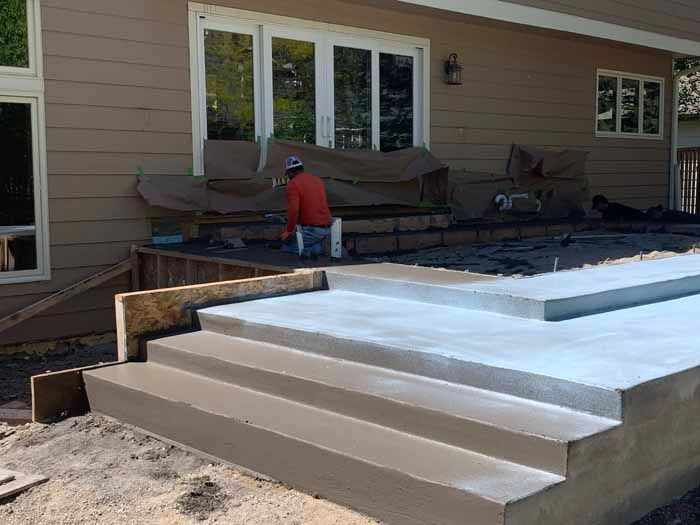 Concrete steps and patio under construction near a house; worker is visible in the background.