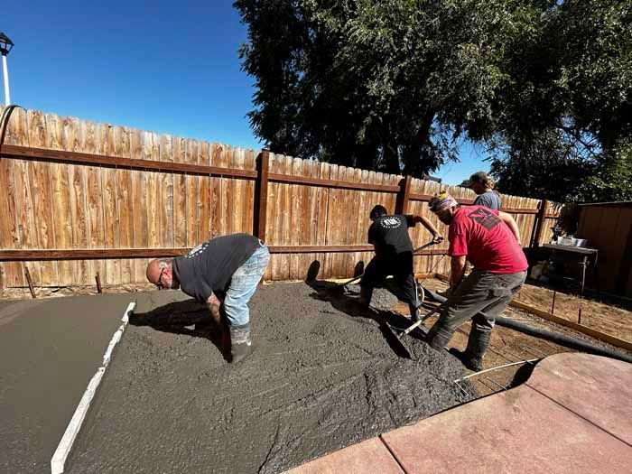 Men working on a concrete patio, spreading and smoothing wet cement near a wooden fence on a sunny day.