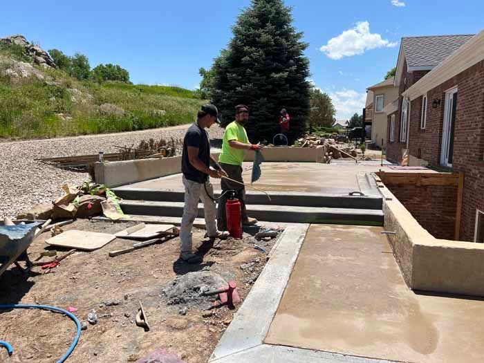 Construction workers on a patio, using tools and equipment outdoors with a building and hill in the background.