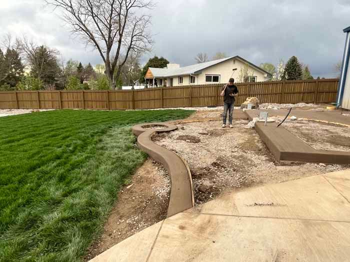 Construction of a concrete border in a backyard with a man standing nearby. Freshly poured concrete, green grass, and wooden fence.
