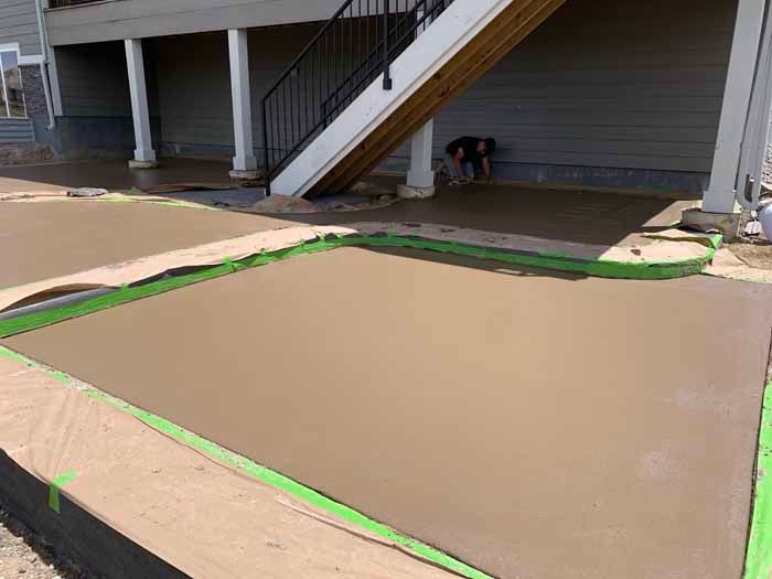 Freshly poured concrete patio with green tape borders; a person works nearby under a deck.