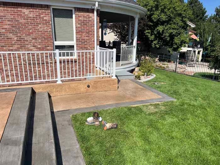 Brick house with white railing and steps leading to a concrete patio, bordered by green grass.