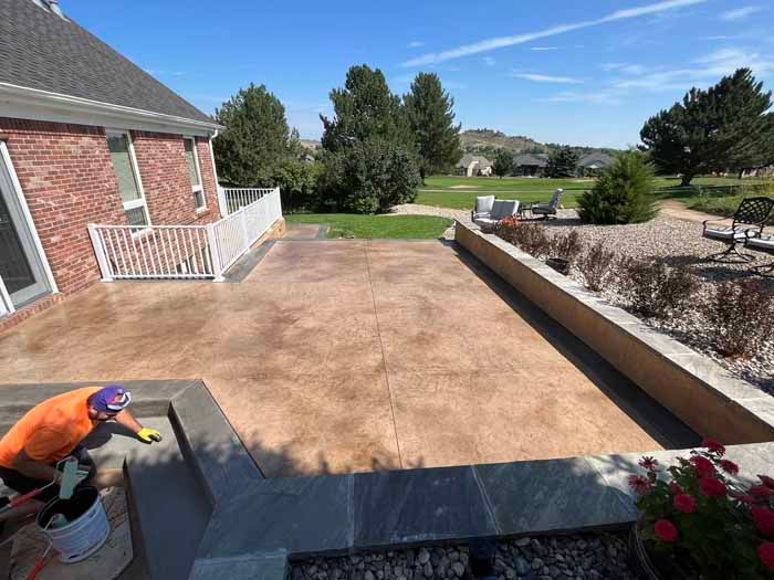 Person staining a large brown patio next to a red brick house, blue sky.
