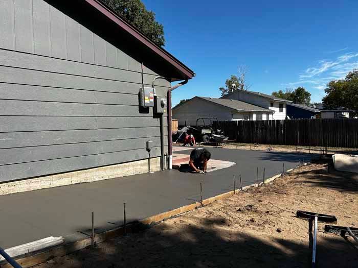Concrete patio being finished next to a gray house, sunny day. Workers, tools, and a wooden form are visible.