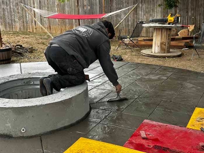 Man troweling wet concrete around a fire pit in a backyard, using gray and black colors.