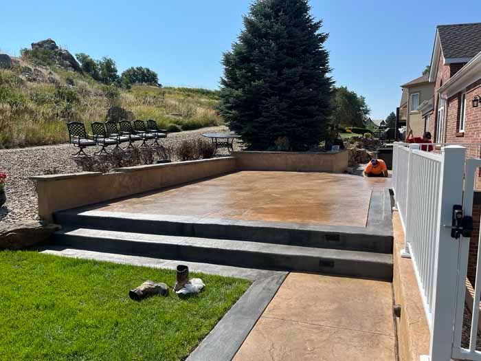 A concrete patio with steps, bordered by a wall, grass, and a white fence. A person works on the patio.