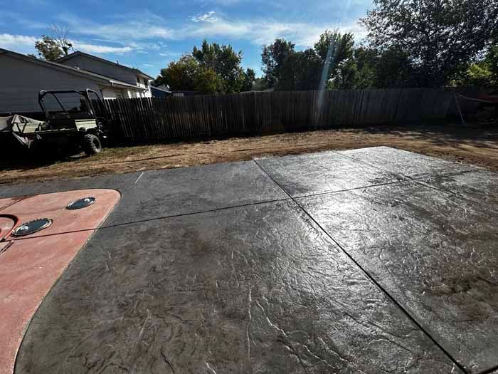 Stamped concrete patio with a dark grey stone pattern. Adjacent dirt area, fence, and utility vehicle visible.
