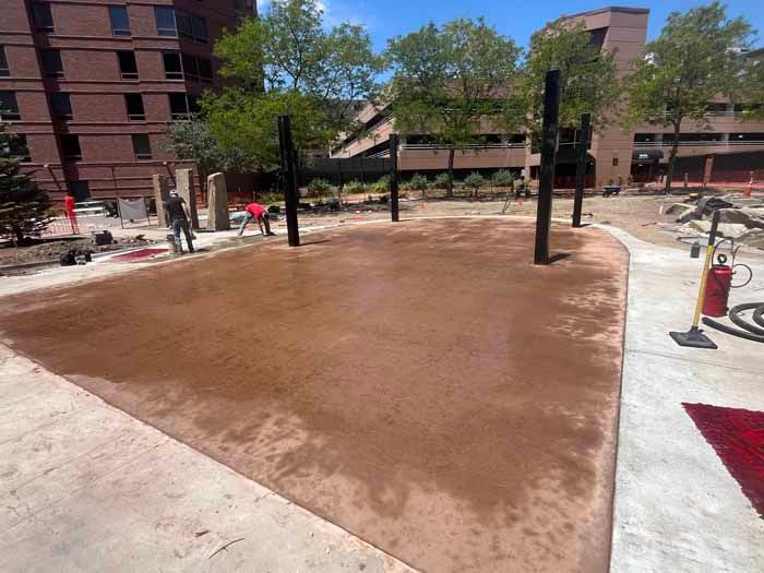 Construction site with wet, reddish-brown concrete square. Workers in red shirts. Black poles and a red rug are in view.