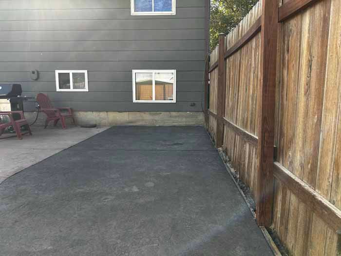 A gray asphalt patio next to a wooden fence and a dark gray house with windows.