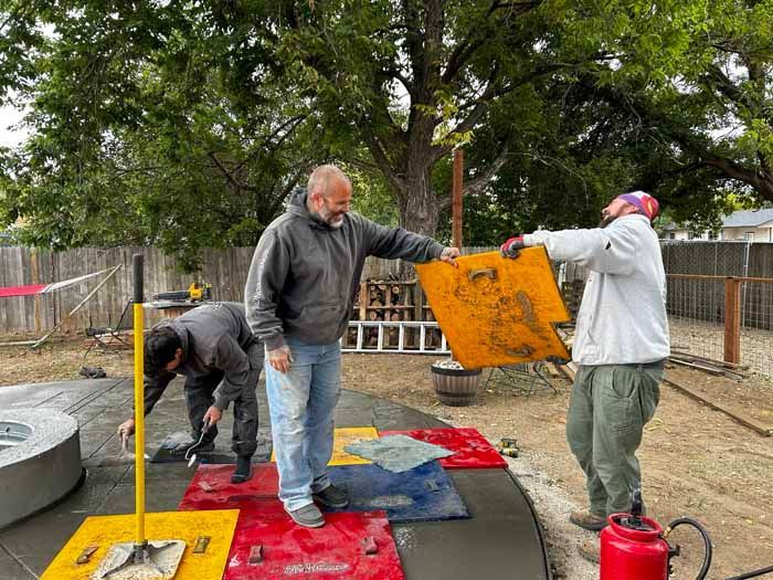 Three people placing colorful concrete pavers in a yard. One holds a yellow mat. Others work nearby.