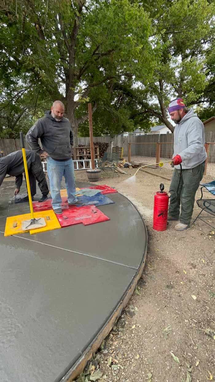 Workers pouring concrete on a curved surface. One holds a fire extinguisher, others use tools. Outdoors, overcast day.