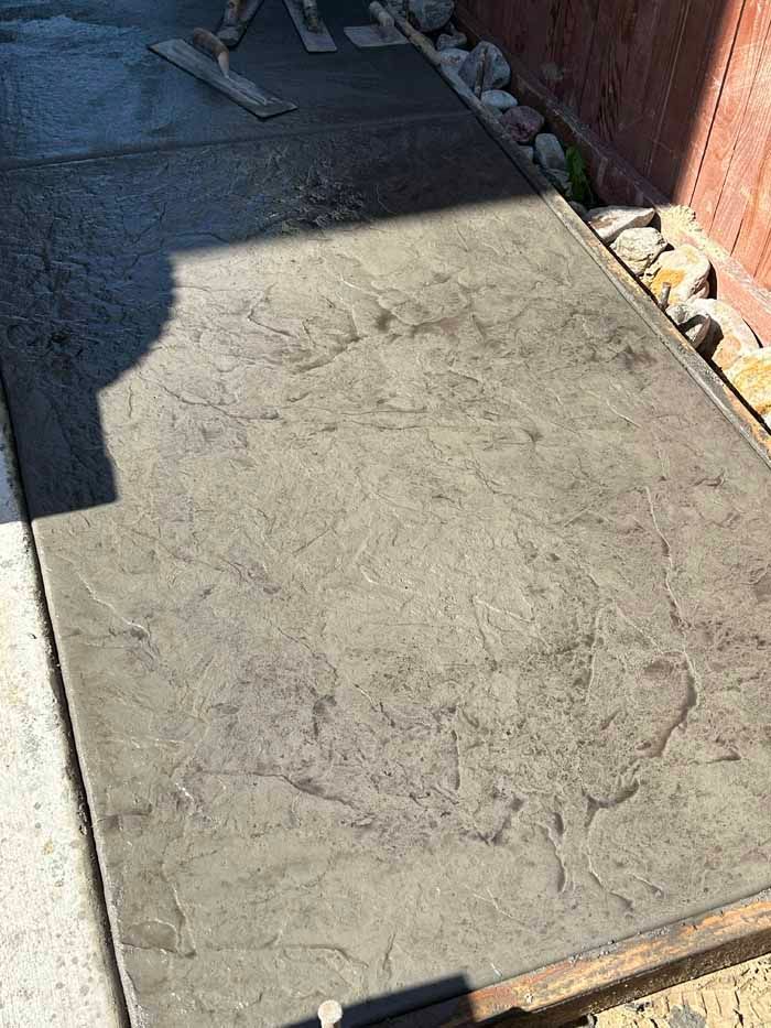 Stamped concrete sidewalk with mottled gray-brown finish, fresh concrete being smoothed in the background.