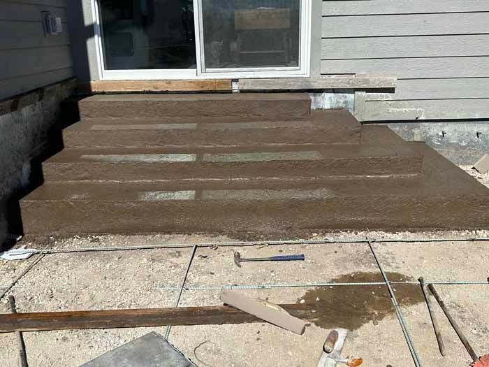 Concrete steps leading up to a sliding glass door; newly poured and wet; set in gravel.