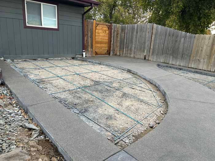 Concrete driveway under construction with rebar grid. Gray concrete borders gravel and wooden fence.