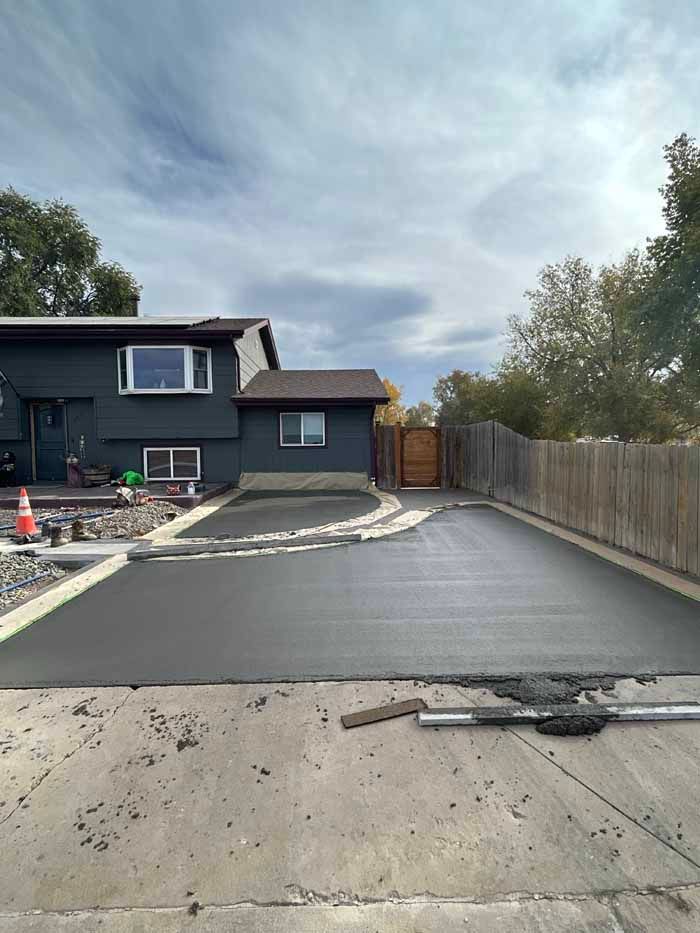 Newly paved asphalt driveway in front of a dark-colored house and wooden fence under a cloudy sky.