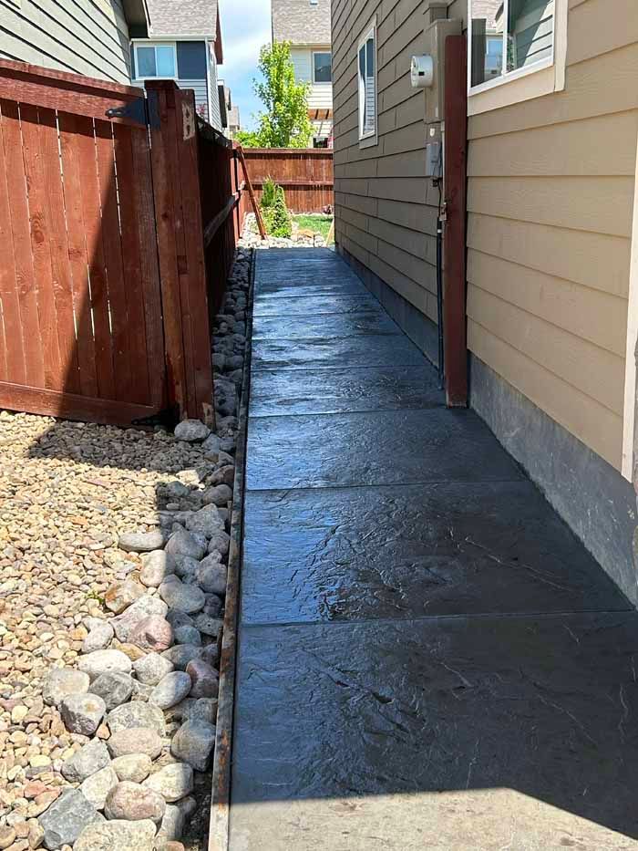 A narrow, dark gray concrete walkway between a wooden fence and a house, with rocks along the fence.
