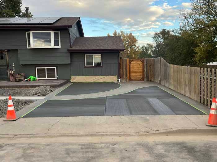 Dark house with gray driveway. Construction cones flank a newly paved section in front of a wooden gate.