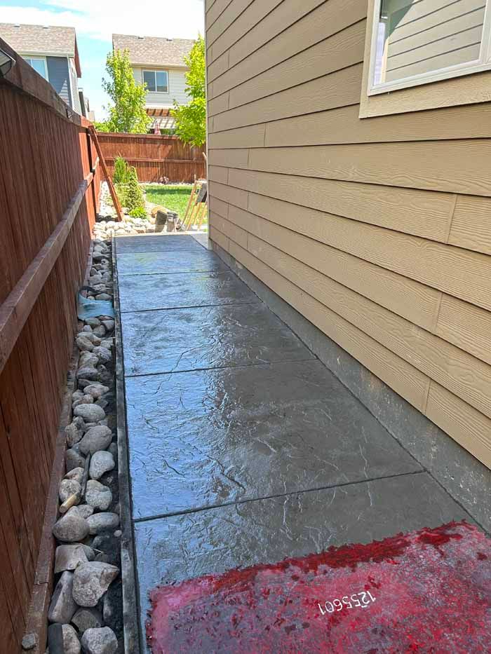 Concrete walkway next to a tan house and a wooden fence; rocks line the fence.