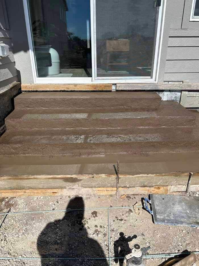 Concrete steps under construction leading to a sliding glass door. A person's shadow is visible.