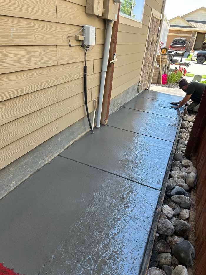 Man smoothing wet concrete sidewalk next to a house; rocks border the edge.